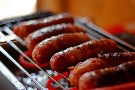 Salchichas asándose en una parrilla junto a rodajas de pimientos y cebollas.