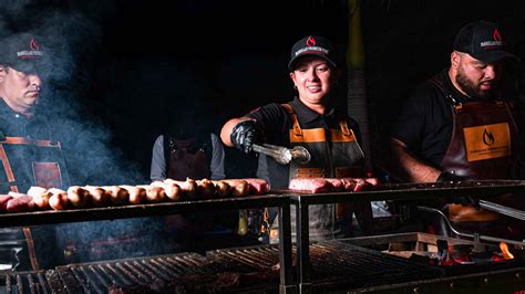Fotografía de un parrillero profesional trabajando con cortes de carne de alta calidad sobre una parrilla encendida.