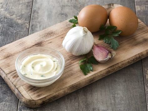 Fotografía gastronómica de un bol con alioli casero sobre una mesa de madera con ajos frescos y perejil alrededor.