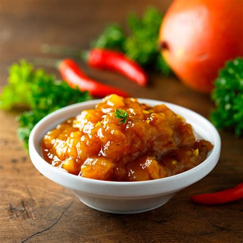 Fotografía gastronómica de un tarro de chutney de mango artesanal acompañado de especias frescas como cardamomo y chiles.