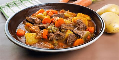 Fotografía de un estofado de jabalí servido en una cazuela de barro, acompañado de patatas doradas y una hogaza de pan.