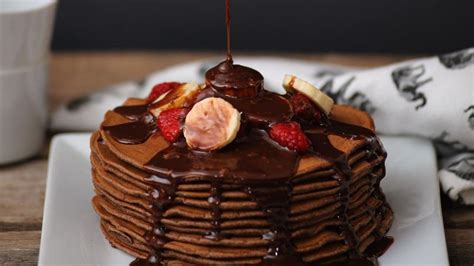Ilustración apetitosa de panqueques de chocolate apilados con salsa de chocolate y frutas frescas.