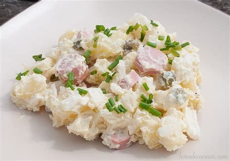 Plato de ensalada alemana de patata (Kartoffelsalat) adornada con hierbas frescas y rodajas de pepinillo, acompañada de salchichas.