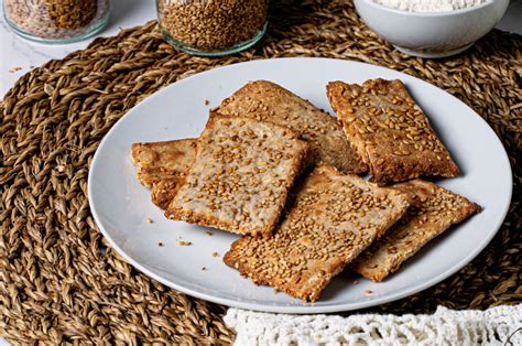 Imagen de una bandeja de horno con crackers de semillas recién horneados, finos y dorados, acompañados de un bol de hummus y vegetales.