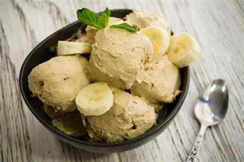 Imagen de un helado cremoso de plátano y manjar servido en un pocillo, decorado con láminas de plátano, líneas de manjar y galletas trituradas.