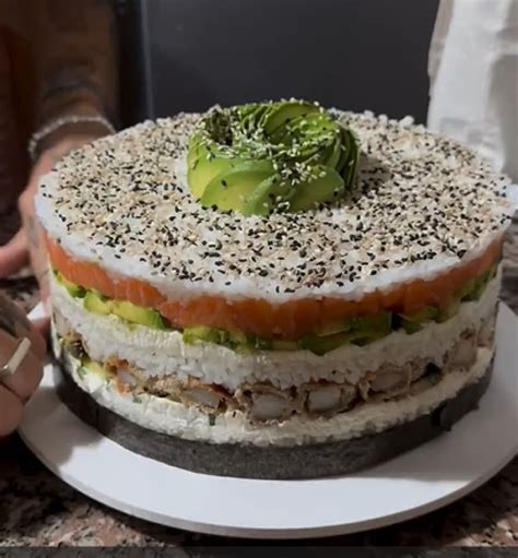 Fotografía detallada de una torta de sushi montada, mostrando las capas de arroz, palta, queso crema y camarones en un aro de emplatado.