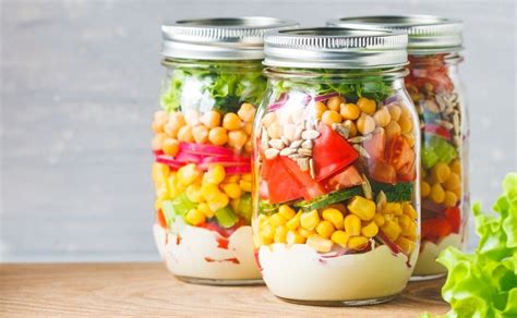 Infografía con las capas de una ensalada en bote: aliño en la base, seguido de legumbres/cereales, verduras densas y hojas verdes en la parte superior.