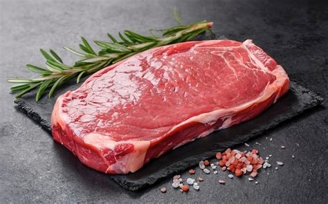 Carne cruda de res (bistec o medallón de filete) marinando en un recipiente con salsa inglesa y otros ingredientes, con hierbas aromáticas visibles.