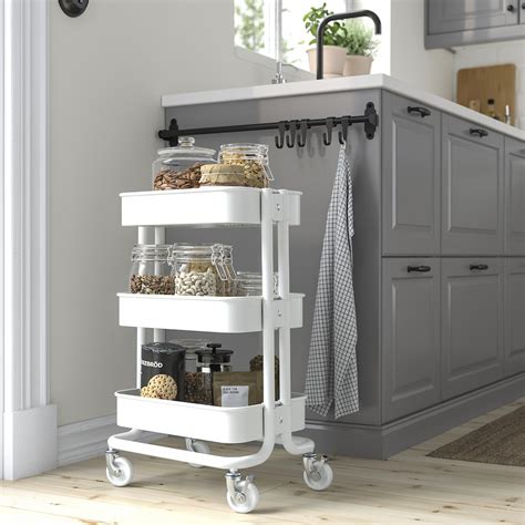 Foto de un carrito verdulero RÅSKOG blanco en una cocina, con verduras frescas y especias en sus cestas