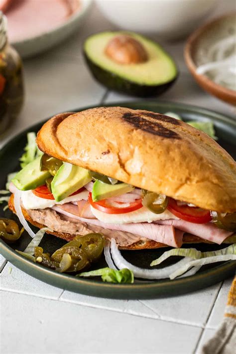 Fotografía de una torta de jamón clásica montada sobre una mesa, mostrando las capas de lechuga, jitomate, aguacate y el pan telera tostado.