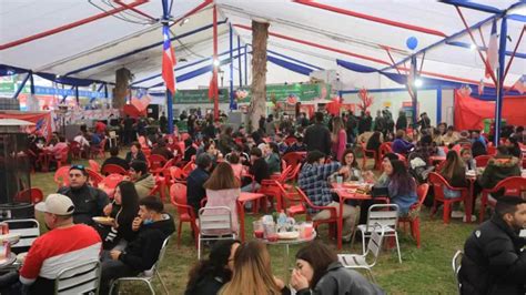 Fotografía panorámica de una fonda chilena en plena celebración, con gente bailando cueca, puestos de comida y parrillas humeantes.