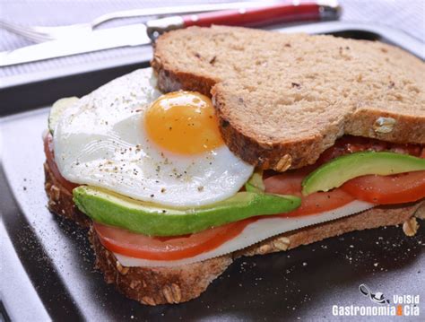 Diagrama paso a paso del montaje de capas del sándwich: pan, puré de aguacate, tomate, queso y mezcla de huevo con mayonesa