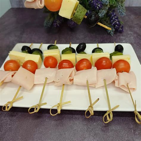 Fotografía profesional de una bandeja con diversas variedades de mini brochetas de queso y jamón sobre una mesa elegante.