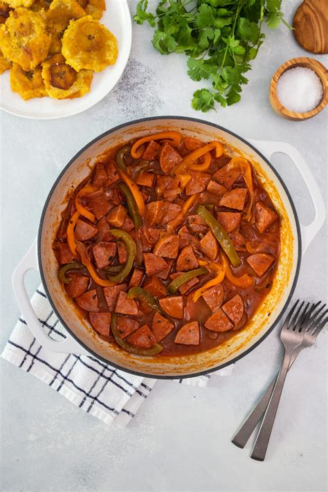 Foto de un plato de salami guisado dominicano humeante con salsa roja espesa, servido junto a mangú de plátano verde