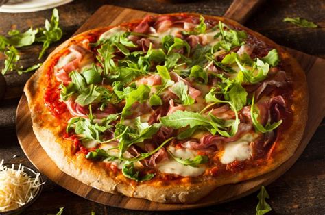 Esquema de ingredientes frescos para pizza: jamón serrano, rúcula, tomates cherry y parmesano dispuestos sobre una mesa
