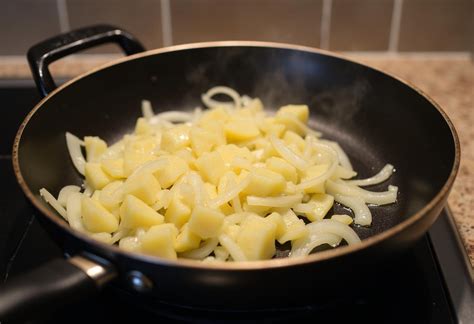Primer plano de patatas y cebolla pochándose lentamente en una sartén con aceite de oliva.