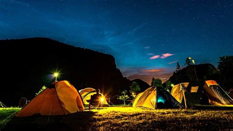 Diseñador: Una foto nocturna de un campamento urbano acogedor, con tiendas de campaña decoradas con luces LED, gente alrededor de una fogata simulada y pasteles listos para comer.