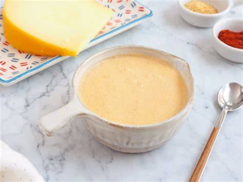 Fotografía gastronómica de una salsa de queso cremosa en un bol rodeada de trozos de queso variado y una cuchara de madera