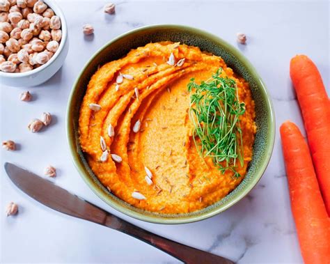 Imagen de un bol de hummus de zanahoria decorado con aceite de oliva, pimentón y brotes frescos junto a bastoncitos de pan.