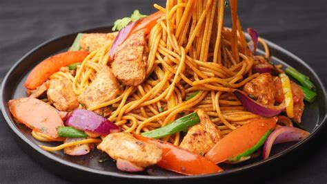 Un wok lleno de tallarín saltado con fideos, trozos de pollo y vegetales coloridos salteándose a alta temperatura.