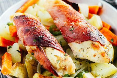 Fotografía gastronómica de pechugas de pollo envueltas en jamón serrano sobre una cama de vegetales horneados.